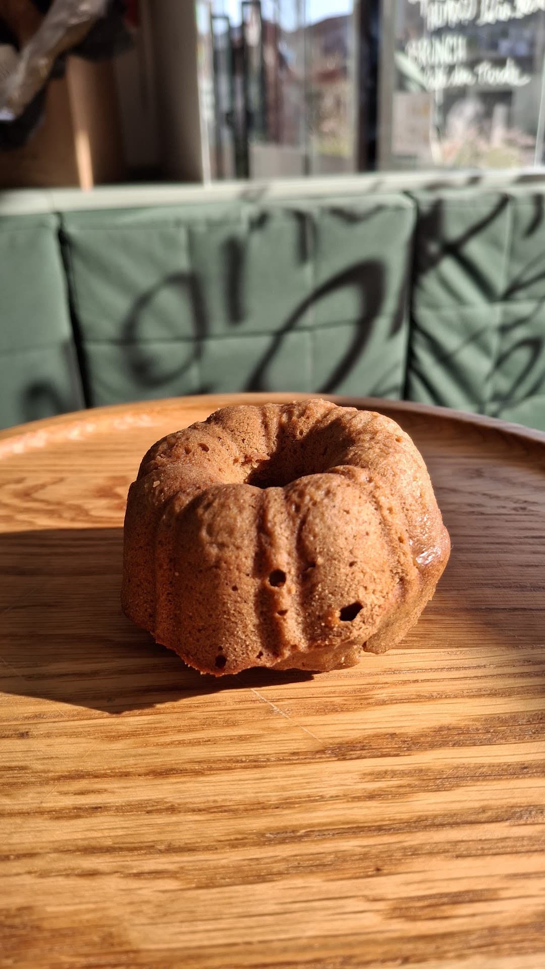 Bolinho artesanal servido em bandeja de madeira clara, banhado pela luz da janela, tendo atrás o banco estofado verde do salão do Zepe com a assinatura da marca estampada e a fachada envidraçada com o letreiro escrito à mão.