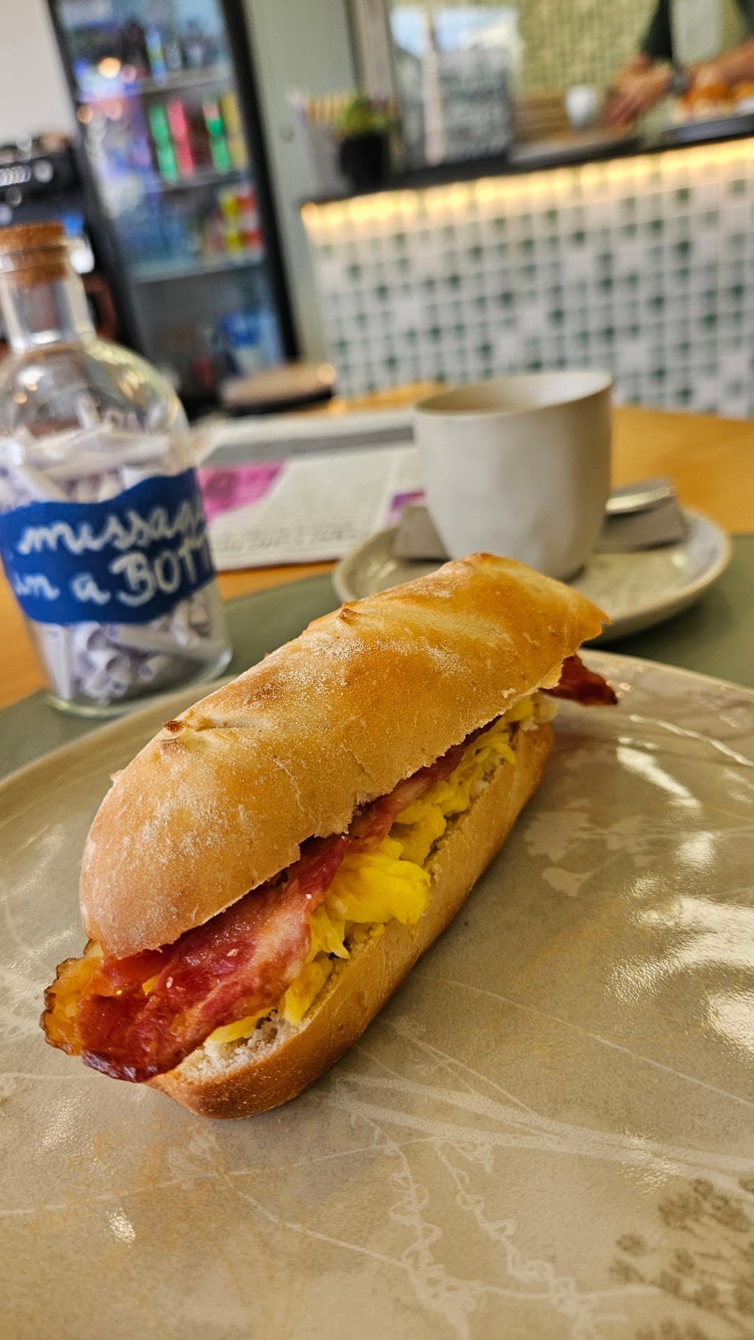 Baguete Italiana recheada com ovos mexidos cremosos e bacon, servida em prato artesanal. Ao lado, o vidro da casa com o rótulo manuscrito "message in a BOTTLE", cheio de bilhetes de papel enrolados, e uma xícara de café atrás.