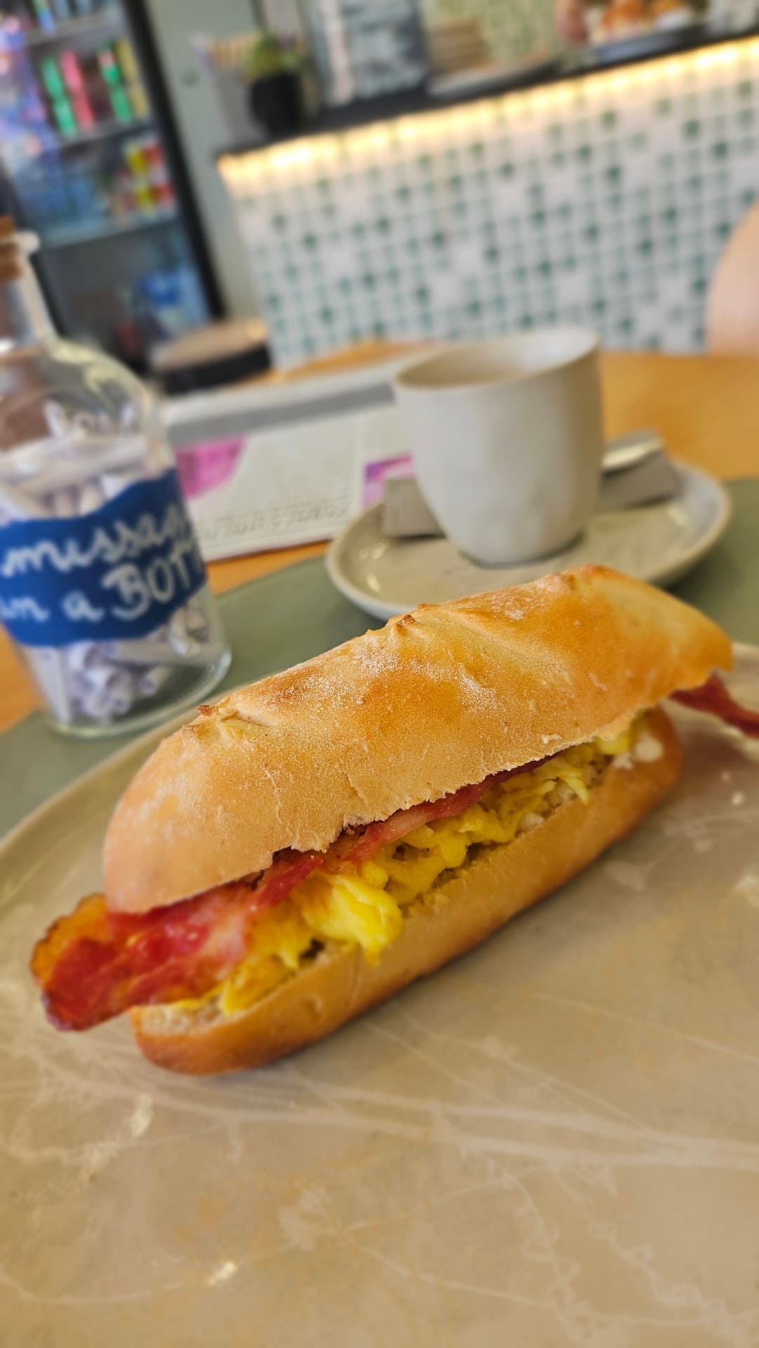 Baguete Italiana com ovos mexidos e bacon em prato artesanal, com o vidro de bilhetes da casa, um exemplar do Jornal do Zepe e uma xícara de café ao fundo, diante do balcão de pastilhas verdes.