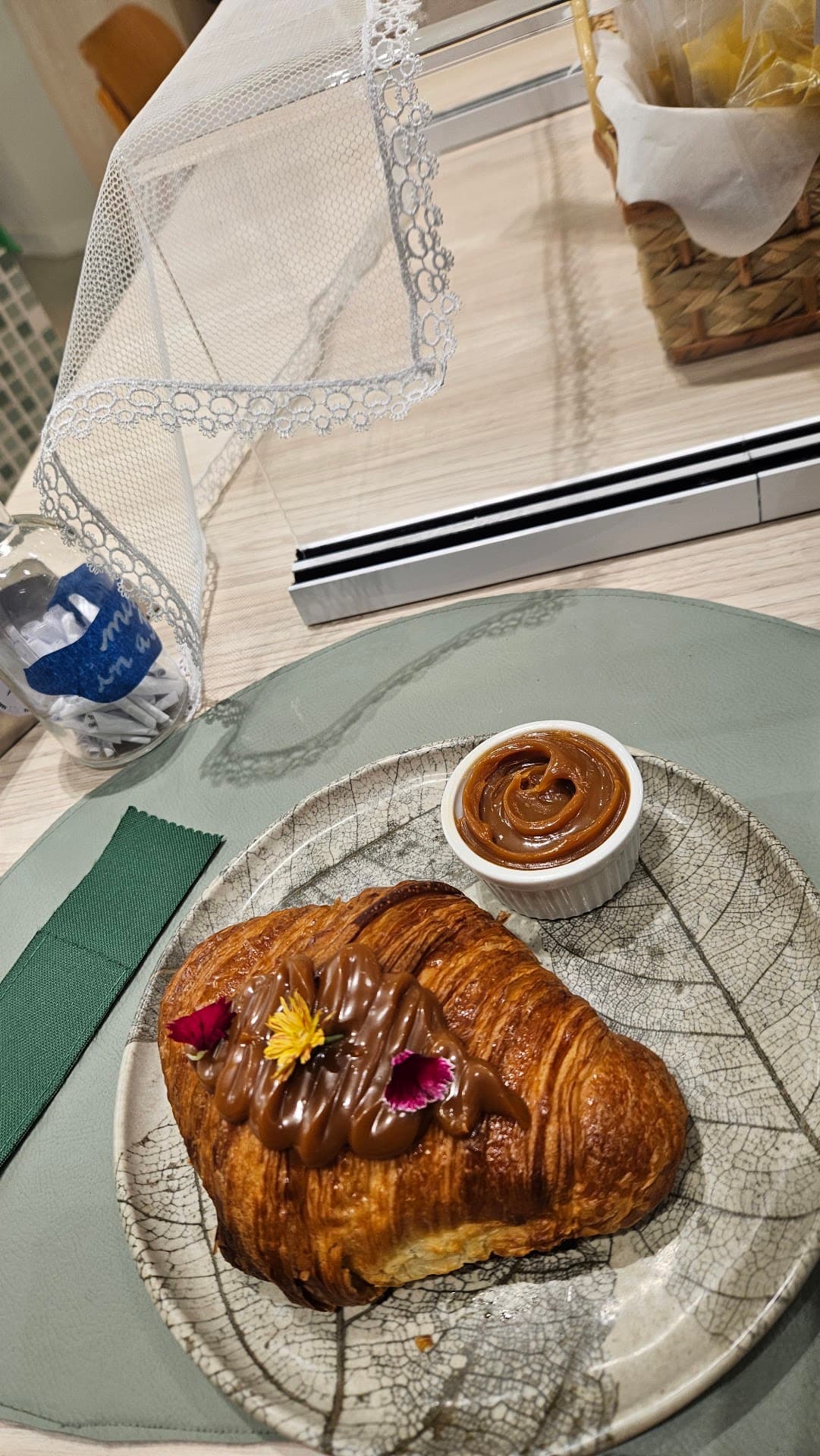 Mesa do Zepe com croissant de doce de leite em prato artesanal, guardanapo verde, e o vidro transparente cheio de bilhetes enrolados ao lado.