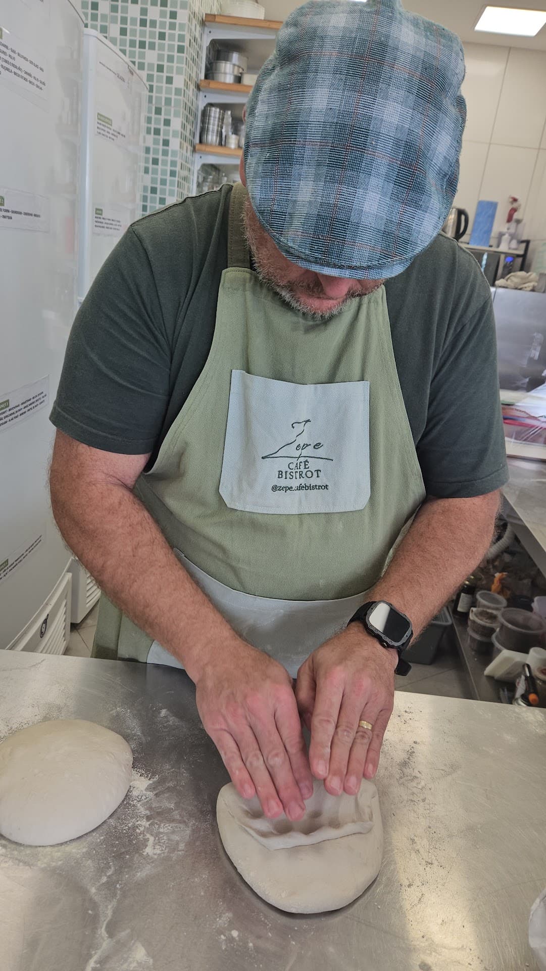 Chef Marcelo, de avental bordado com o logo do Zepe Café Bistrot, abrindo com as duas mãos um disco de massa de fermentação natural sobre o balcão de inox polvilhado de farinha.