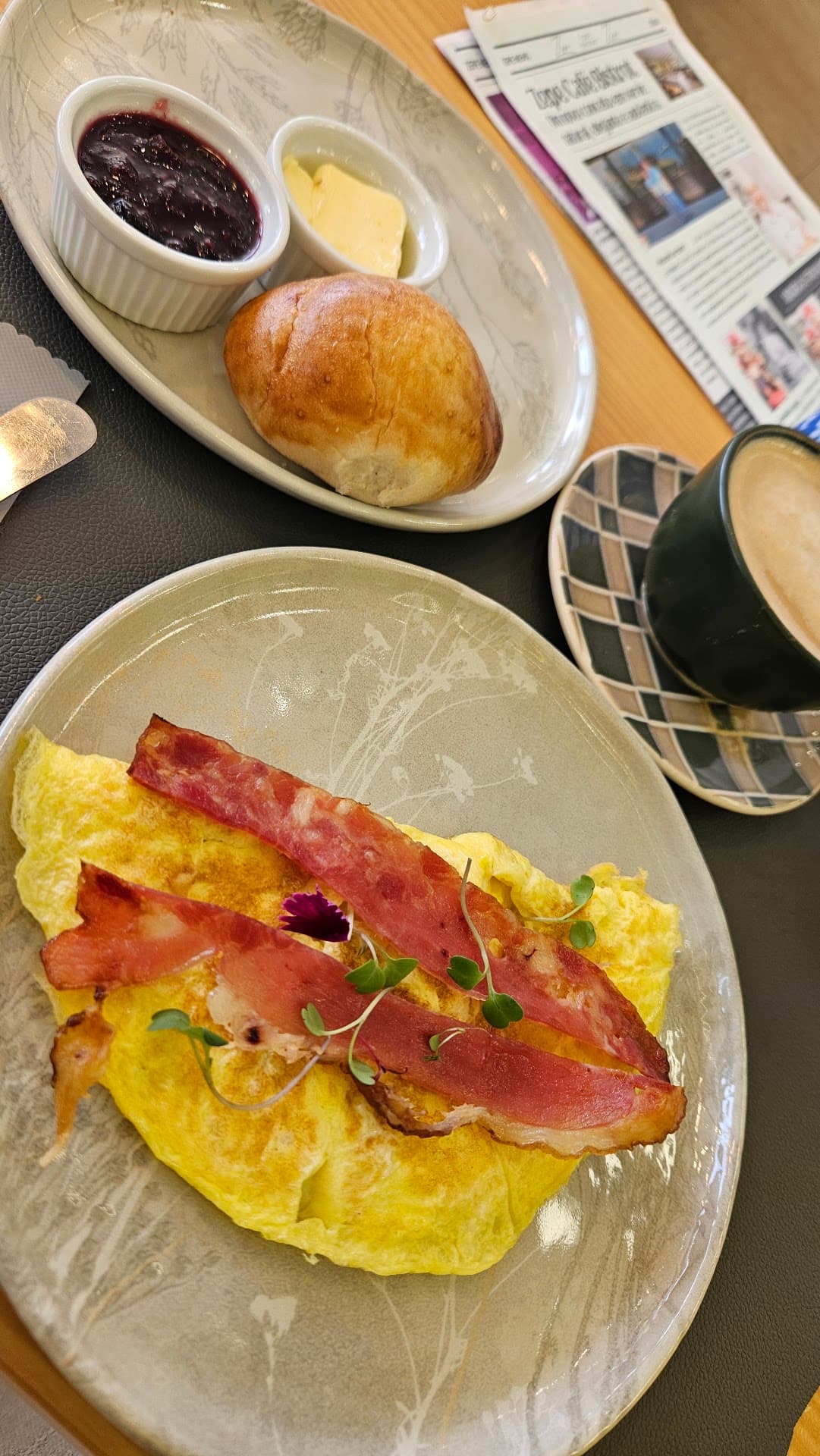 Omelete com fatias de bacon e brotos, pão brioche, manteiga e geleia de frutas vermelhas servidos em pratos artesanais, com um cappuccino ao lado e um exemplar do Jornal do Zepe aberto sobre a mesa.