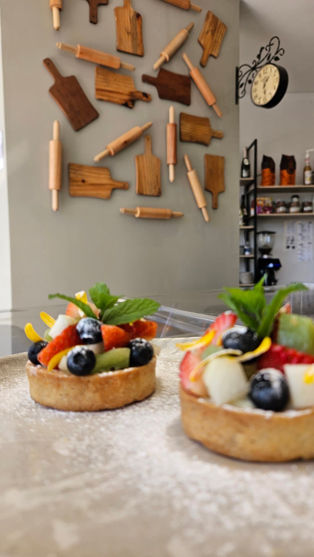 Parede do salão do Zepe coberta por dezenas de rolos de macarrão e tábuas de madeira de tamanhos diferentes, com um relógio de parede antigo à direita e duas tarteletes de frutas em primeiro plano.